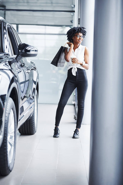 Young african american woman in glasses stands in car salon near vehicle with package in hands. - Φωτογραφία, εικόνα