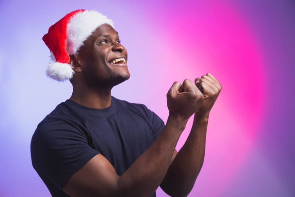 Portrét pozitivního afroamerického usmívajícího se muže v Santa klobouku a ležérním tričku na barevném pozadí, vánoční čas. Zimní prázdniny a koncept emocionálních lidí. - Fotografie, Obrázek