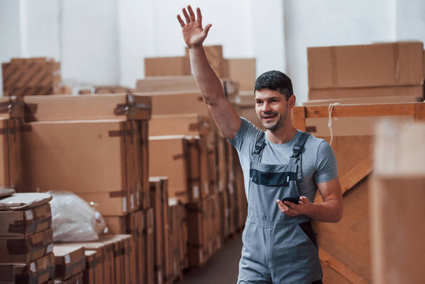 Young storage worker in uniform with phone stands and raised hand up. - Fotoğraf, Görsel