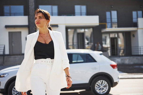 Pewna siebie kobieta w białym, formalnym stroju outdoots w pobliżu jej białego samochodu. - Zdjęcie, obraz