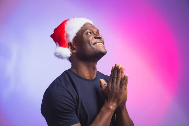 Retrato de homem sorrindo afro-americano positivo em chapéu de Papai Noel e camiseta casual em fundo colorido, tempo de Natal. Férias de inverno e conceito de pessoas emocionais. - Foto, Imagem