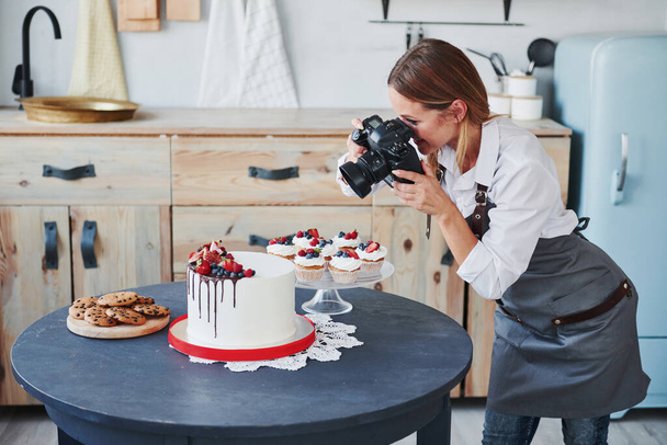 Woman stands in the kitchen and takes photo of her homemade cookies and pie by using camera. - Foto, Imagem