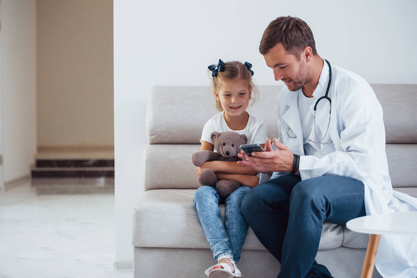 Male doctor in white uniform sits in the clinic with little girl. - Φωτογραφία, εικόνα
