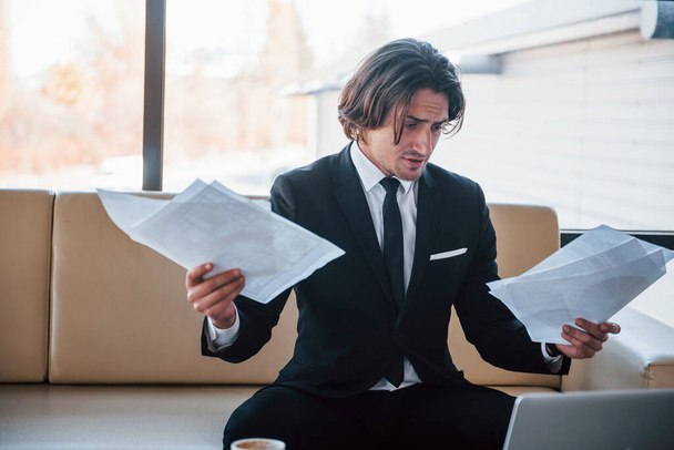 Holds documents in hands. Portrait of handsome young businessman in black suit and tie. - 写真・画像