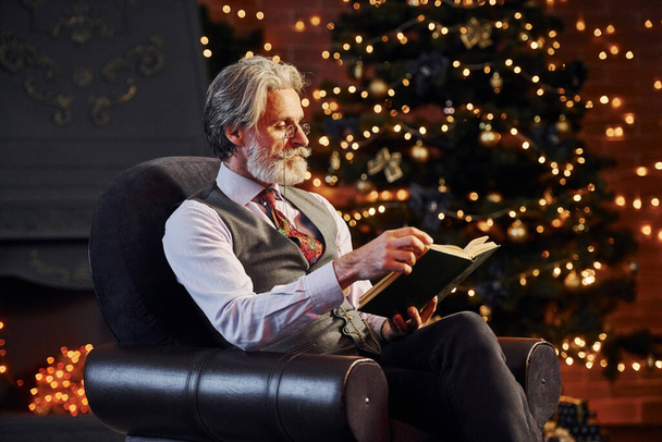 Πορτρέτο της κομψής ανώτερος με γκρι μαλλιά και γενειάδα βιβλίο ανάγνωσης σε διακοσμημένο χριστουγεννιάτικο δωμάτιο. - Φωτογραφία, εικόνα