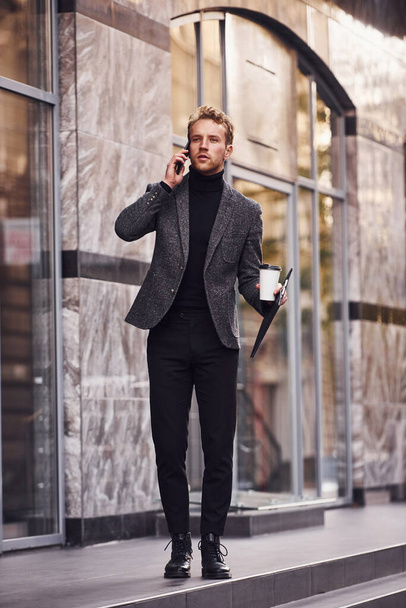 Man in elegant formal wear with cup of drink and notepad in hands is outside against modern building have a conversation by the phone. - Foto, imagen