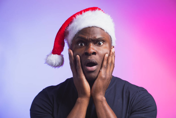 Portrait of surprised african american man in santa hat and casual t-shirt on colourful background, christmas time. Winter holidays and emotional people concept. - Photo, Image