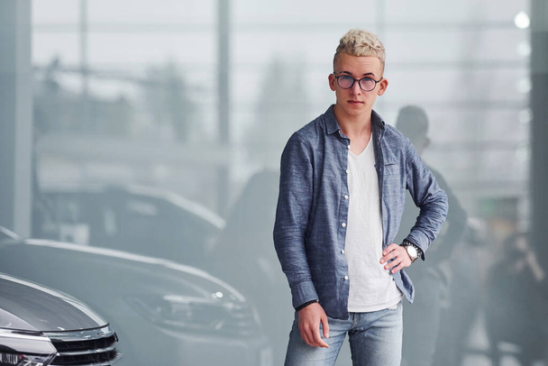 Young hipster guy in nice clothes stands indoors against grey background near modern car. - Foto, Imagem