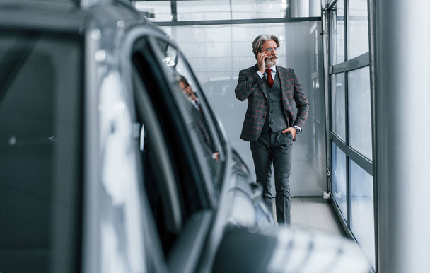 Starší podnikatel v obleku a kravatě s šedými vlasy a vousy stojí uvnitř s telefonem v blízkosti auta. - Fotografie, Obrázek
