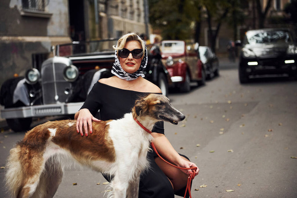 Blonde woman in sunglasses and in black dress near old vintage classic car with her dog. - Фото, изображение