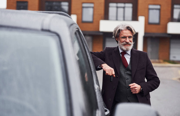 Fashionable senior man with gray hair and beard standing outdoors on the street near his car with keys in hand. - 写真・画像