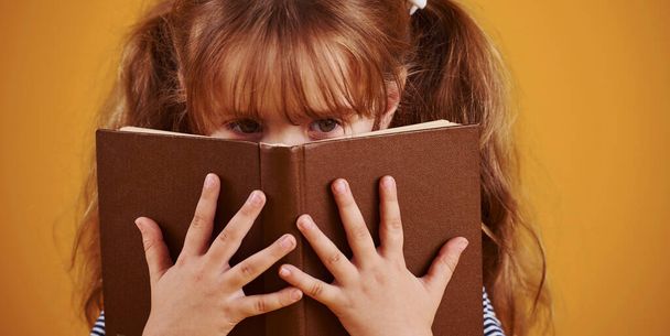 Cute little girl with book in hands in the studio against yellow background. - 写真・画像