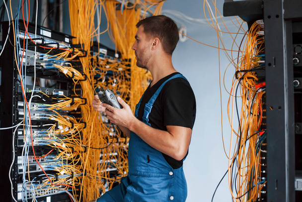 Young man in uniform with measuring device works with internet equipment and wires in server room. - Foto, Bild