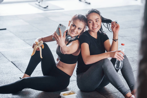 Two young girls in sportive clothes sitting with food in the gym together at daytime and making a selfie. - Фото, зображення