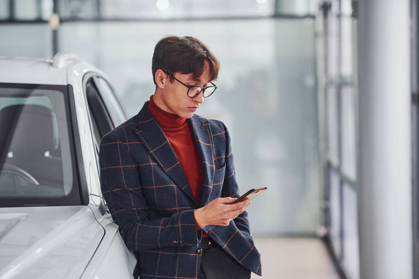 Hipster in formal wear and in eyewear standing near white modern car indoors. - Photo, Image