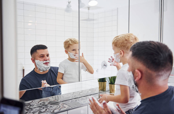 Father with his son is in the bathroom have fun by using shaving gel and looking in the mirror. - Foto, imagen