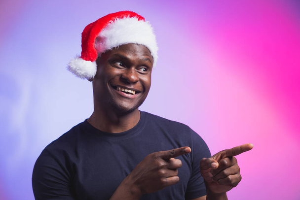 Portrait d'un homme souriant afro-américain positif en chapeau de Père Noël et t-shirt décontracté sur fond coloré, période de Noël. Vacances d'hiver et les gens émotionnels concept. - Photo, image