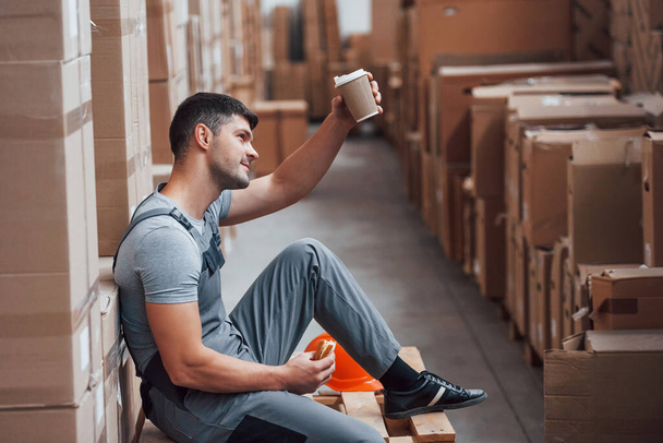 Storage worker sits and have a break. Eats sandwitch and drinks coffee. - Foto, imagen
