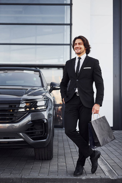 Portrait of handsome young businessman in black suit and tie outdoors near modern car and with shopping bags. - Fotografie, Obrázek