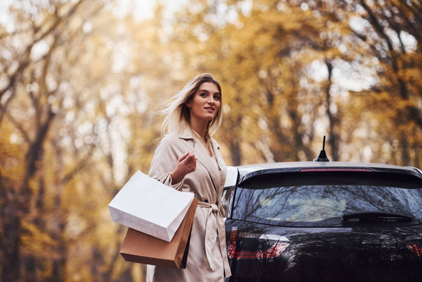 Girl walks near the car with shopping bags in hands. Modern brand new automobile in the forest. - Fotoğraf, Görsel