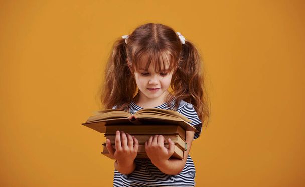 Cute little girl with book in hands in the studio against yellow background. - Foto, afbeelding