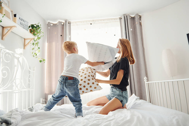 Mother playing pillow fight with her son in bedroom at daytime. - 写真・画像