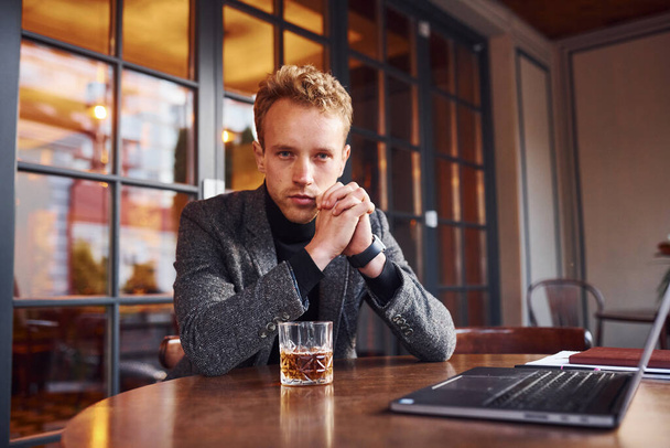 Resmi kıyafetli zarif bir genç, kafeteryada dizüstü bilgisayarı ve bir bardak alkolle oturuyor.. - Fotoğraf, Görsel