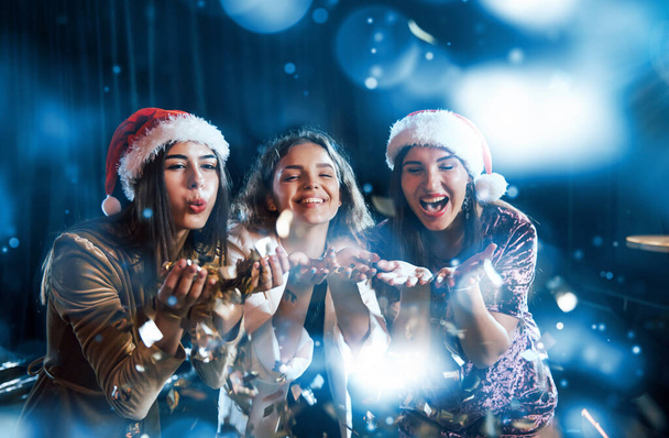 Three women blowing confetti from their hands indoors at holiday time. - Photo, Image