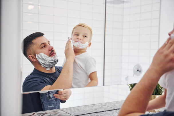 Father with his son is in the bathroom have fun by using shaving gel and looking in the mirror. - Foto, imagen