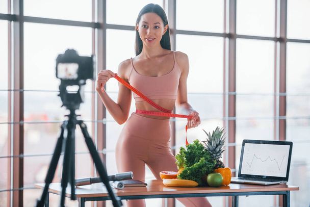 Young fitness vlogger doing video indoors by standing near table with healthy food and laptop. - Фото, изображение