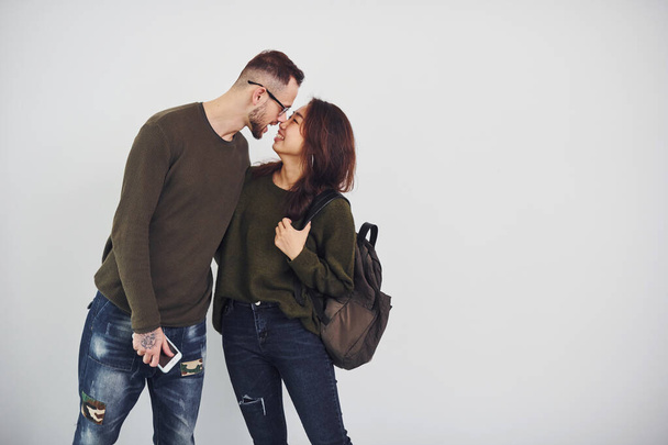 Cheerful multi ethnic couple with backpack and phone standing together indoors in the studio against white background. - 写真・画像