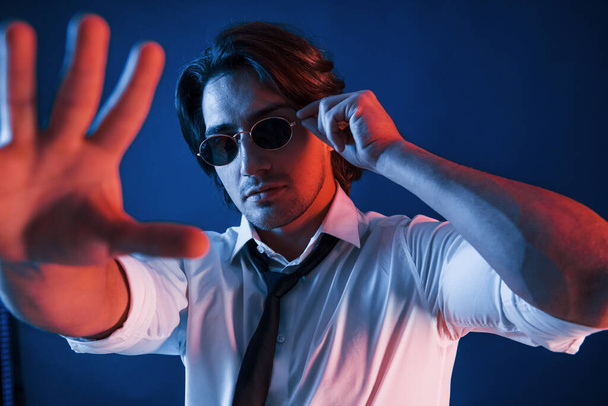 Beautiful man in glasses is in the studio with blue neon lighting holds hand in front of him. - 写真・画像
