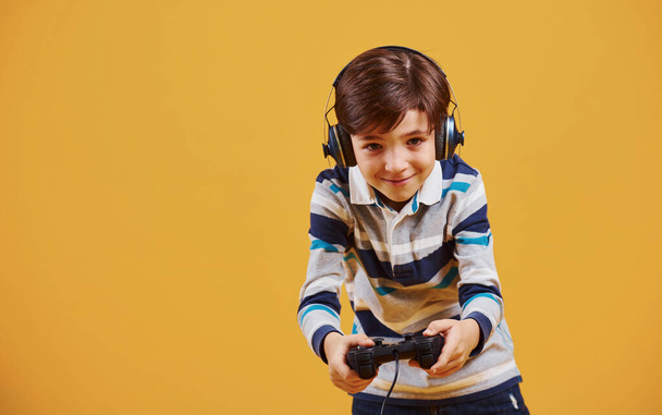 Cute little boy playing video game in the studio against yellow background. - Φωτογραφία, εικόνα