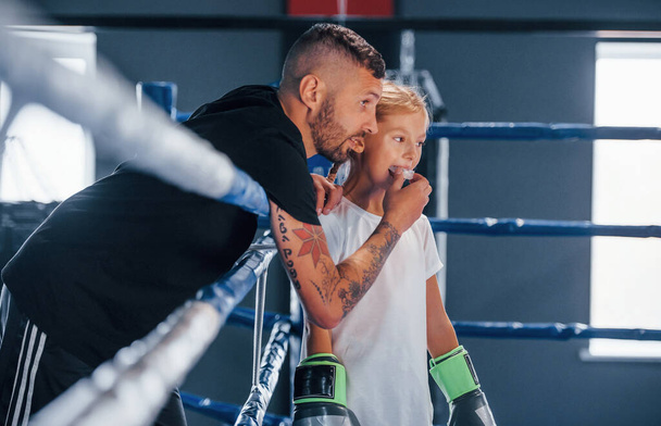 Standing on the boxing ring. Young tattooed boxing coach teaches cute little girl in the gym. - 写真・画像