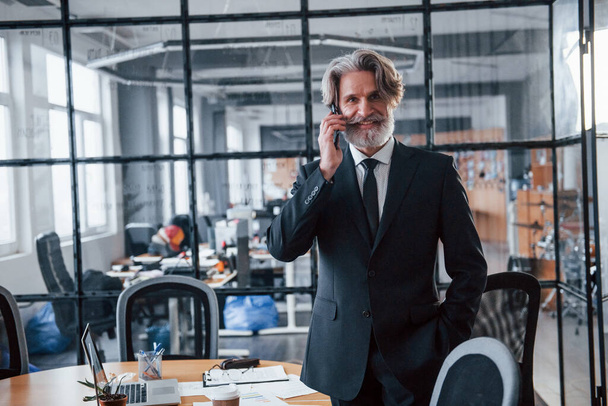 Mature businessman with grey hair and beard in formal clothes have conversation by the phone in the office. - Foto, immagini