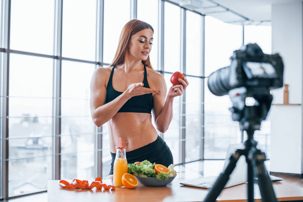 Vrouwelijke vlogger met sportief lichaam dat bij tafel staat met gezond eten. - Foto, afbeelding