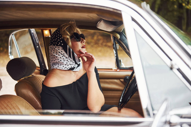 Blonde woman in sunglasses and in black dress sits in old vintage classic car. - Valokuva, kuva