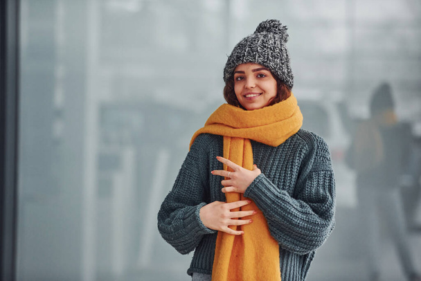 Beautiful cheerful girl in yellow scarf and in warm clothes standing indoors against background with reflections. - Fotografie, Obrázek
