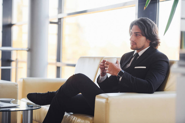 Leül a kanapéra egy csésze itallal. Egy jóképű fiatal üzletember portréja fekete öltönyben és nyakkendőben.. - Fotó, kép