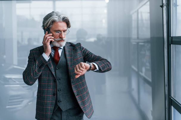 Senior businessman in suit and tie with gray hair and beard standing indoors with phone looking at watch. - Foto, afbeelding