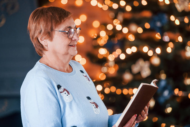 Portrait of grandmother that standing in New year decorated room with book. - Foto, Imagen