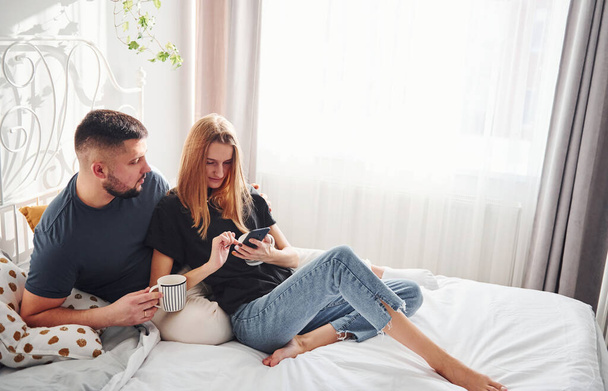 Holding phone and cup of drink. Young married couple have leisure together in bedroom at daytime. - Fotografie, Obrázek