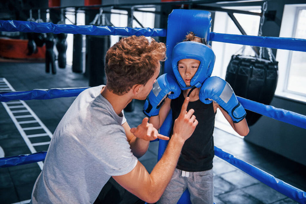 Νεαρός προπονητής πυγμαχίας βοηθά μικρό αγόρι σε προστατευτική ενδυμασία στο ρινγκ μεταξύ των γύρων. - Φωτογραφία, εικόνα