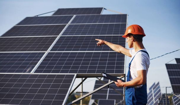 Showing by the hand. Male worker in blue uniform outdoors with solar batteries at sunny day. - Foto, Imagen