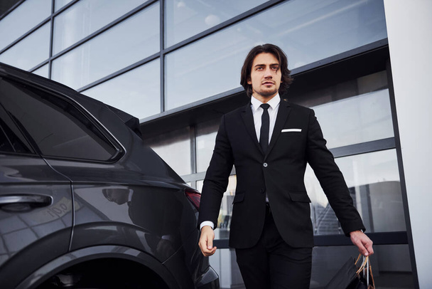 Portrait of handsome young businessman in black suit and tie outdoors near modern car. - Фото, изображение