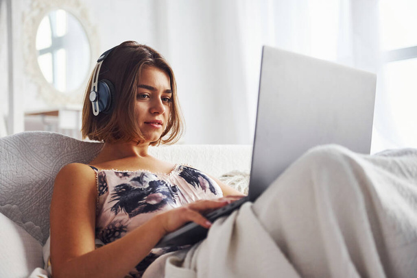 Listening to the music by using headphones. Cute girl lying on the bed with laptop in the bedroom at her weekend time. - Фото, изображение