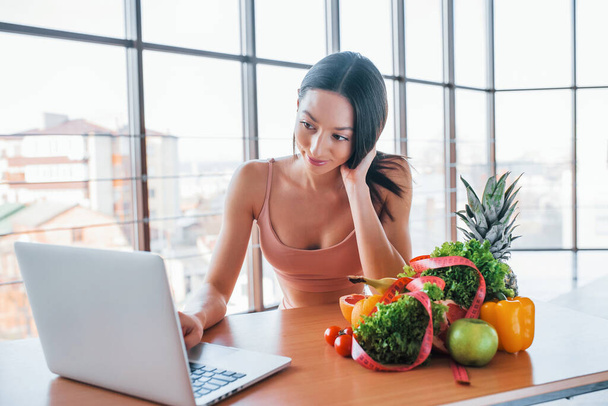 Brunette with slim body type sits by the table with laptop and healthy food. - Foto, Imagen