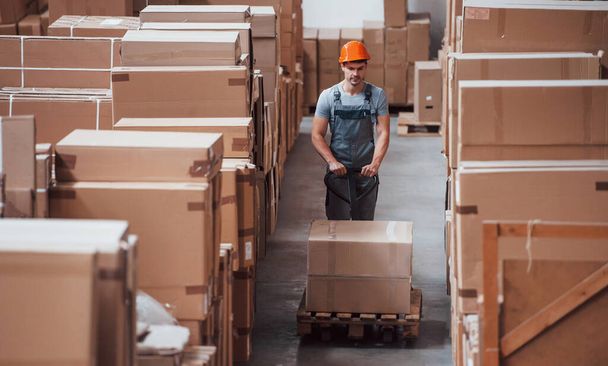 Young male worker in uniform is in the warehouse with pallet truck. - Fotoğraf, Görsel
