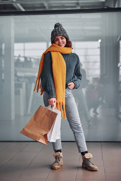 Beautiful cheerful girl in yellow scarf and in warm clothes standing indoors with shopping bags in hands. - Φωτογραφία, εικόνα