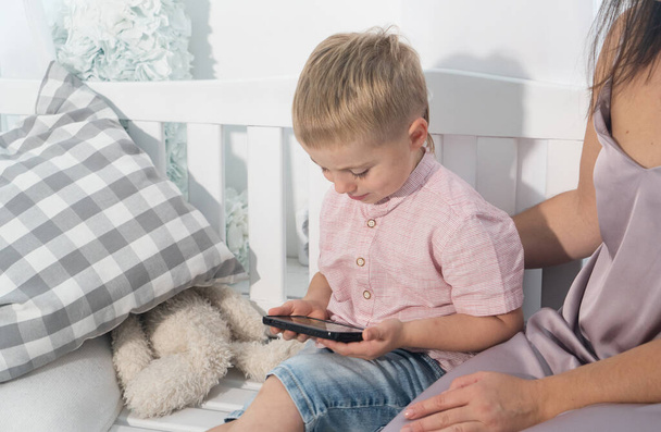 Young woman with her little son sitting on bed and using smartphone - Foto, afbeelding